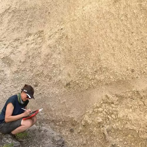 Vicki Smith crouches next to stratigraphy, She is writing notes
